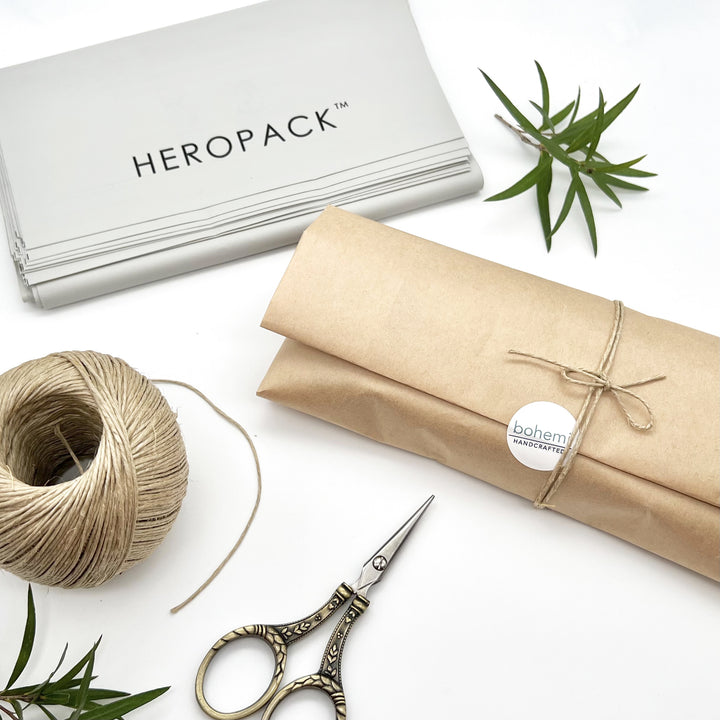 rectangle package wrapped in brown kraft paper with twine bow and sticker reading bohemi, Backround scissors, twine, grey bags reading heropack and green leafy branch.