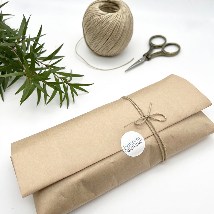 rectangle package wrapped in brown kraft paper with twine bow and sticker reading bohemi, Backround scissors, twine and green leafy branch.